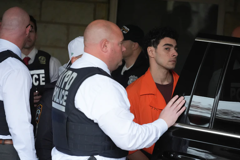 Young man in an orange jumpsuit is escorted into a vehicle by a group of police officers.