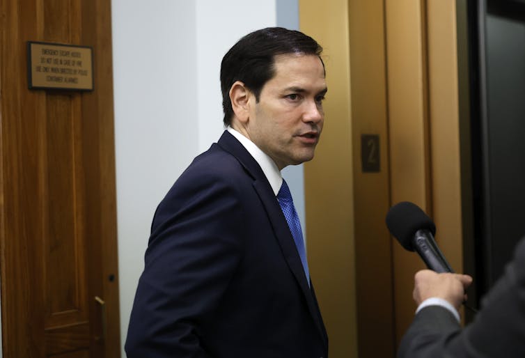 A man with short dark hair and a black blazer and blue tie speaks into a microphone.
