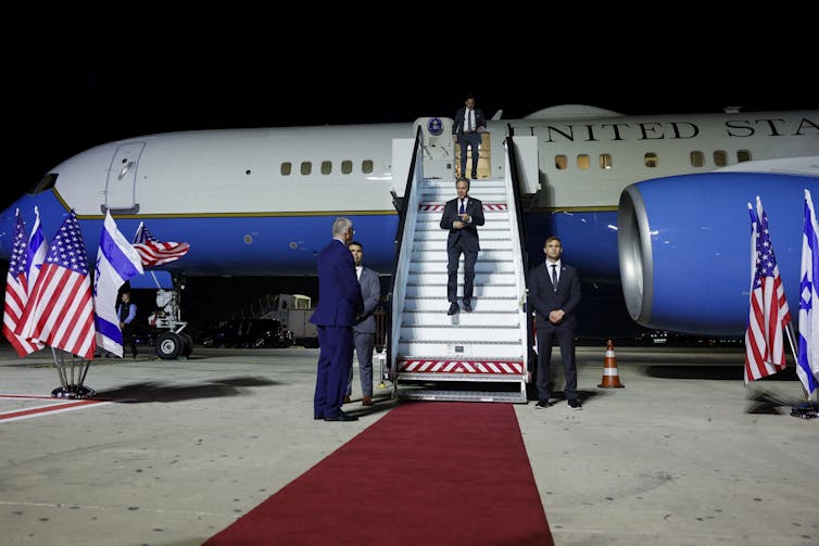 A man wearing a dark suit walks down the steps from a white and blue plane at nighttime. There are several people wearing formal clothing and American flags near the plane.