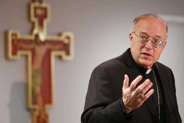 A gray-haired man wearing a traditional black priestly robe stands beside a crucifix mounted on a plain wall.