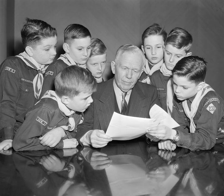An older man with a suit looks at a sheet of paper and is surrounded by a group of young boys wearing Cub Scout uniforms.