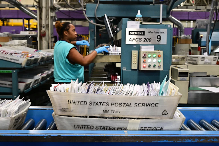 United States Postal Service trays of mail roll by on a conveyor belt in a warehouse. A woman in a turquoise shirt sorts through mail while wearing blue medical gloves.