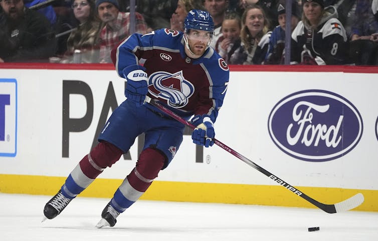 A hockey player in a burgundy and blue uniform skates on an ice rink