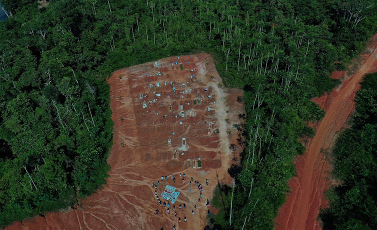 An aerial view of a section of rainforest cut out revealing fresh earth. People gather in the space.