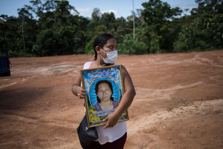 A woman wearing a COVID mask standing in a forest clearing holds a large portrait of a woman.