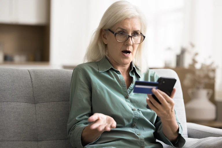 Une femme à l’air choqué tient un téléphone et une carte bancaire.