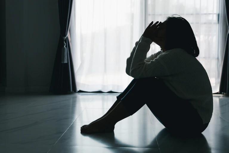 Black and white photo of a silhouette of a young woman sitting on the floor, with her elbows resting on her knees and her hands held up to her face.