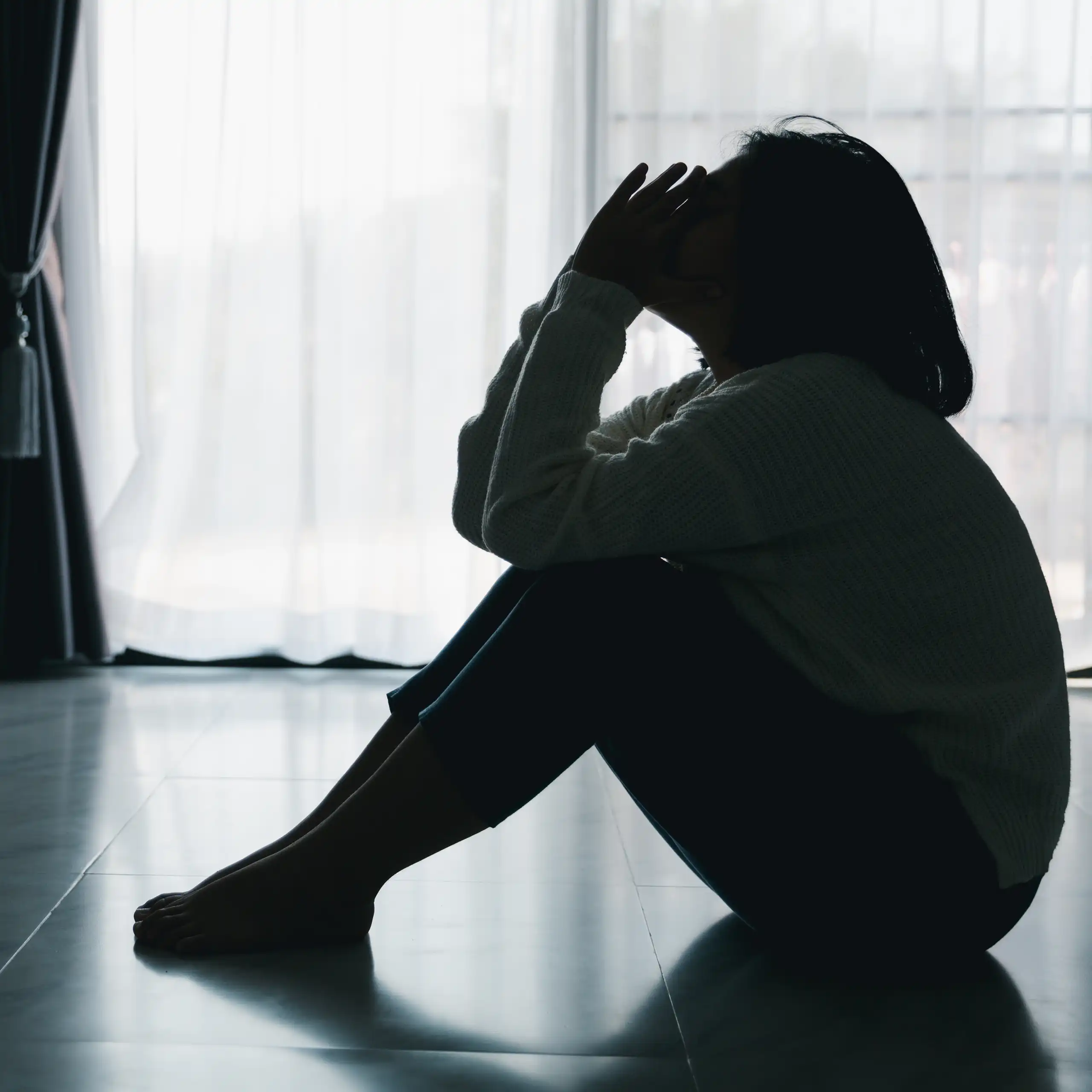 Black and white photo of a silhouette of a young woman sitting on the floor, with her elbows resting on her knees and her hands held up to her face.