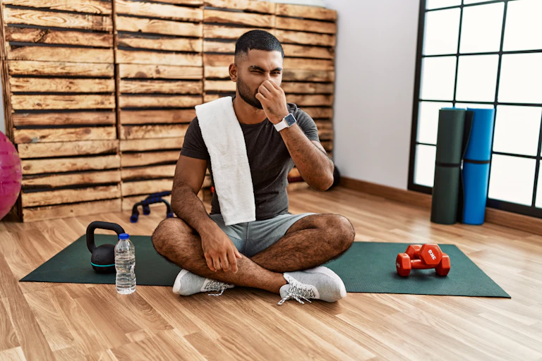 A man sits on an exercise mat in the gym, plugging his nose due to a bad smell.