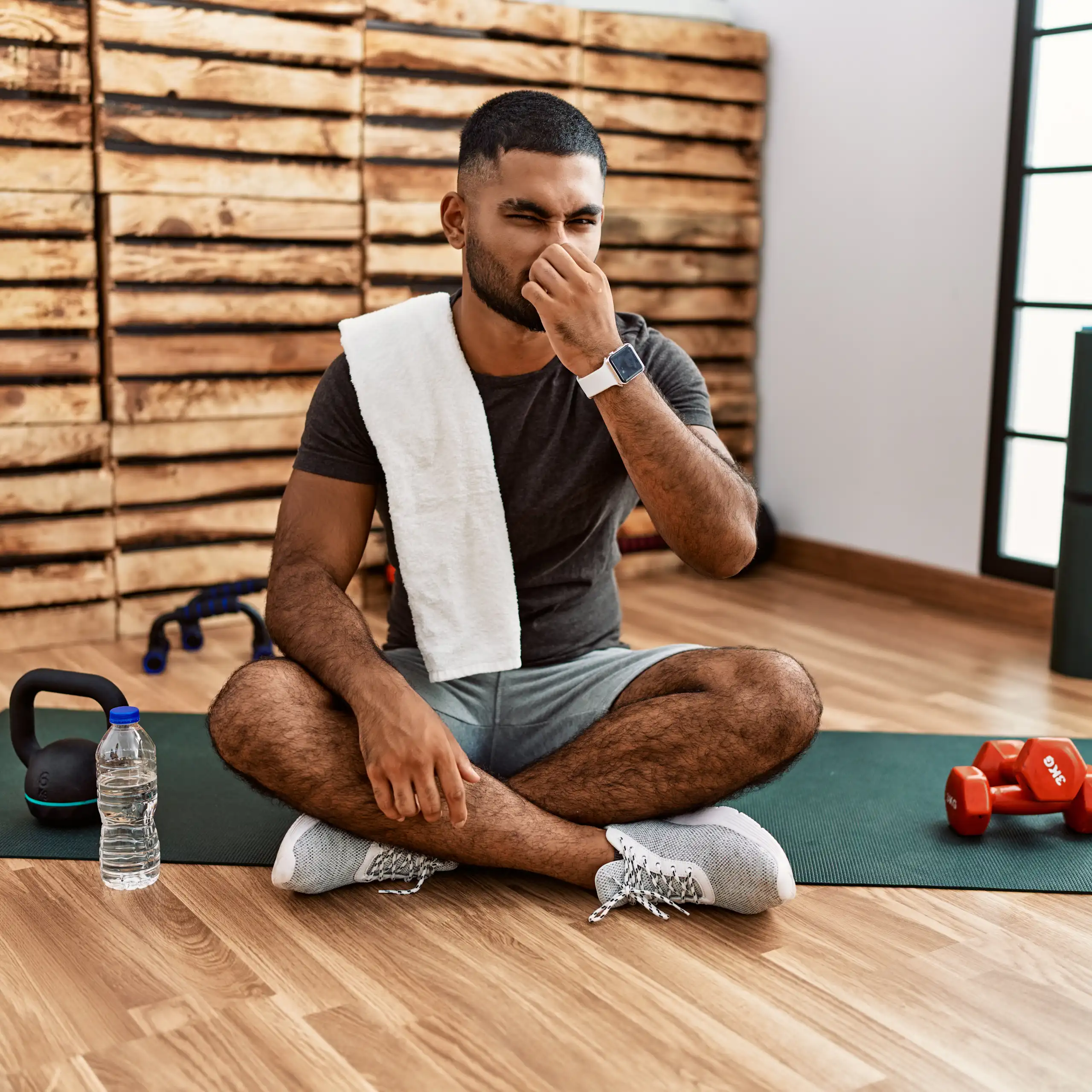 A man sits on an exercise mat in the gym, plugging his nose due to a bad smell.