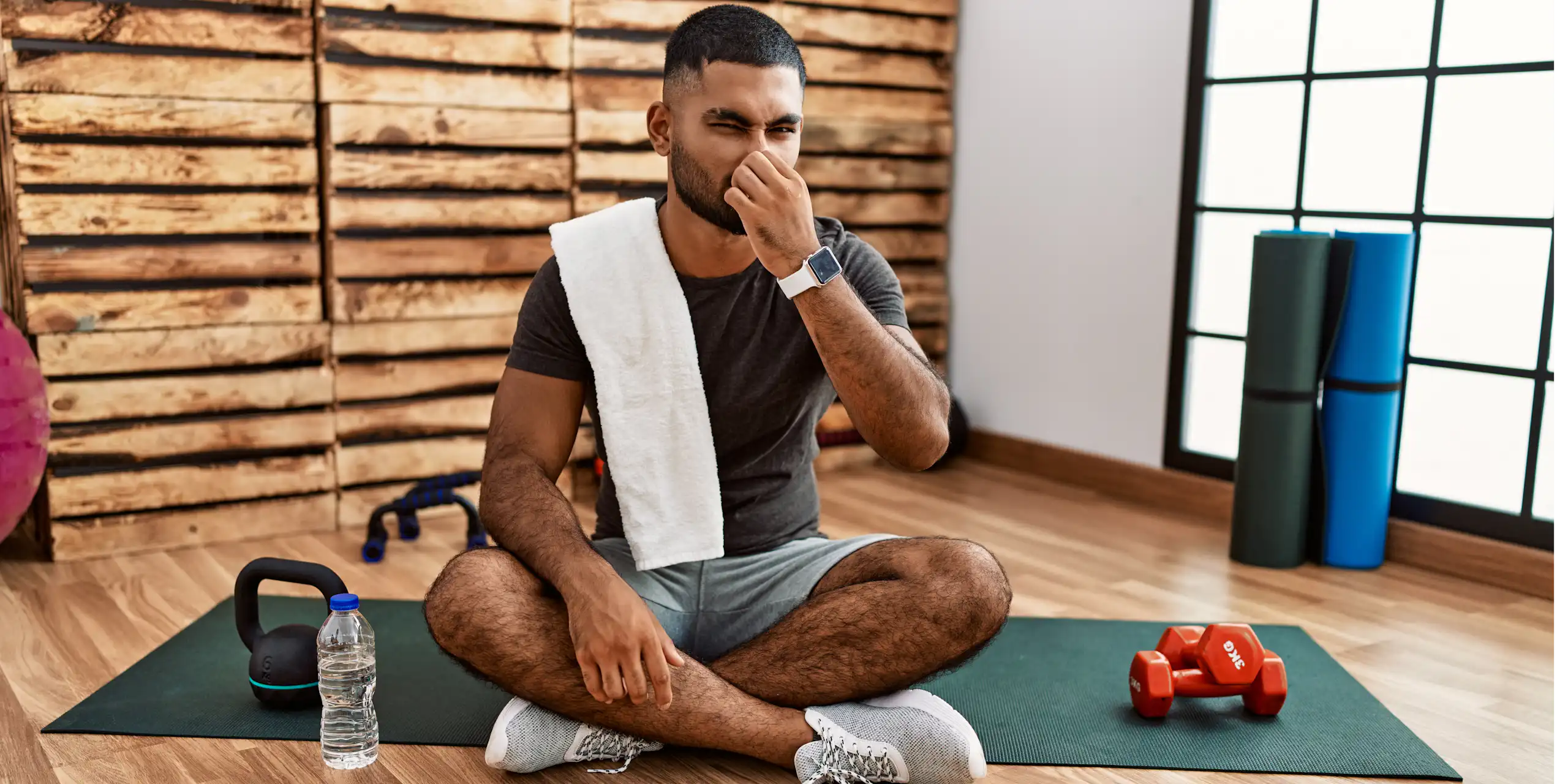 A man sits on an exercise mat in the gym, plugging his nose due to a bad smell.