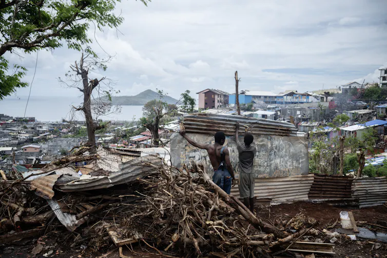 deux garçons de dos face à un paysage dévasté à Mayotte soulèvent un morceau de tôle