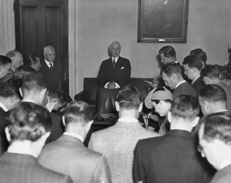 A man in a suit sitting at the front of a room while others stand opposite him.