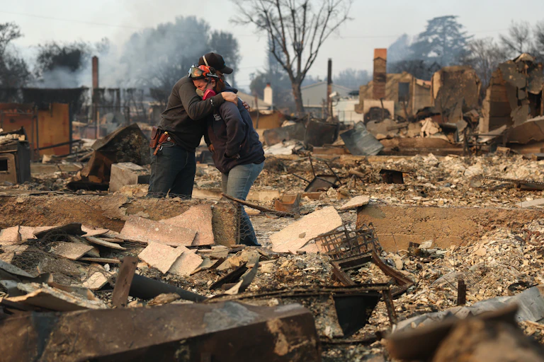 Two people wearing masks embrace amid charred rubble.