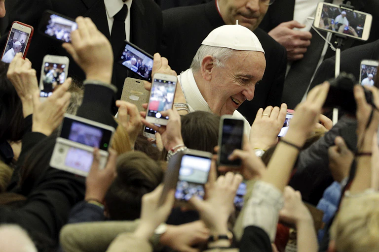 An elderly man in a white skullcap passes through a crowd of people holding up phones to take pictures of him.