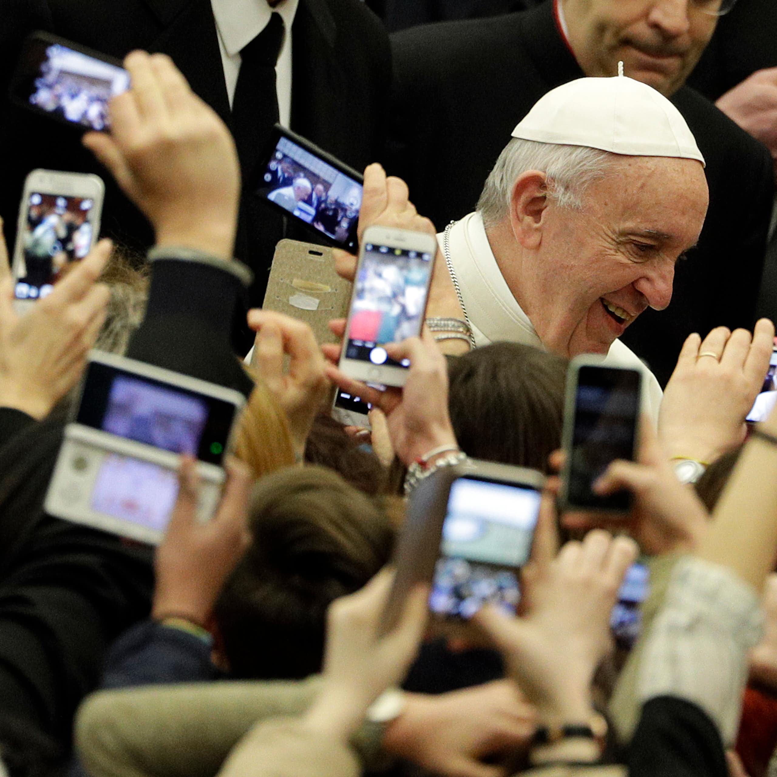 An elderly man in a white skullcap passes through a crowd of people holding up phones to take pictures of him.