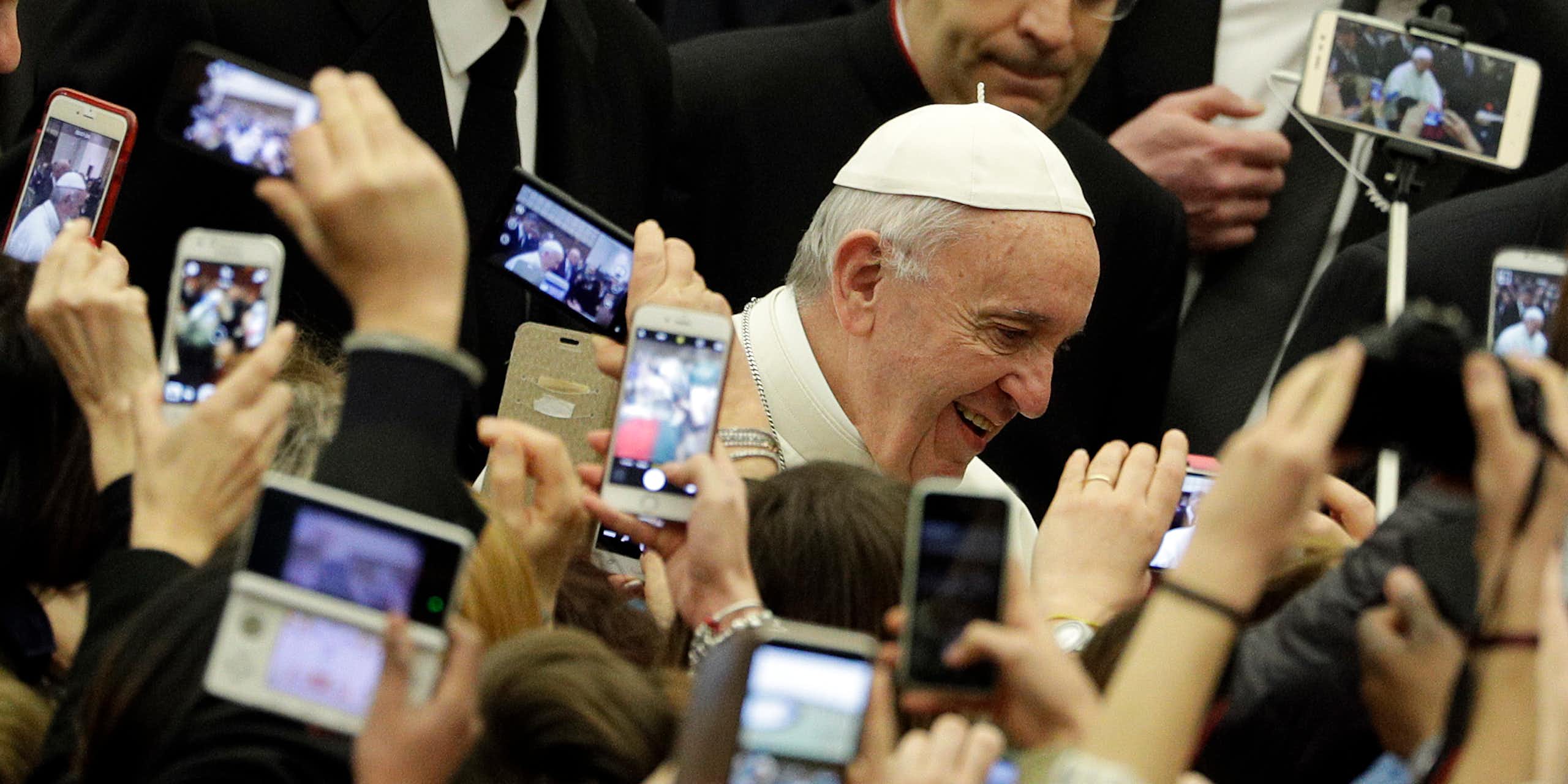 An elderly man in a white skullcap passes through a crowd of people holding up phones to take pictures of him.