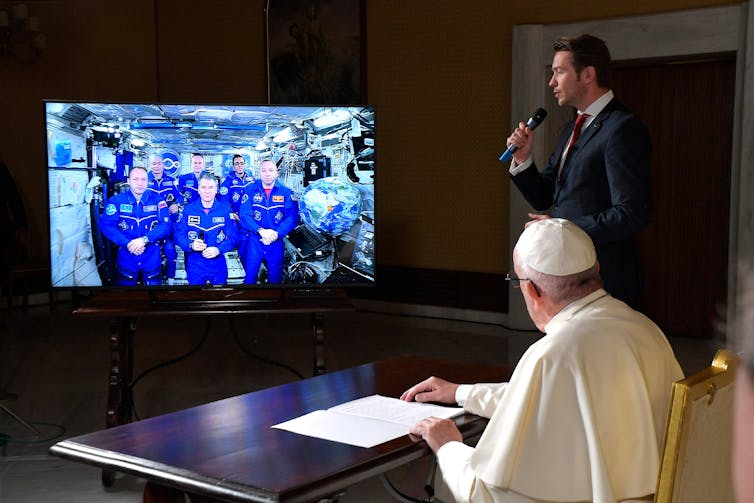A man in white robes sits at a table facing a large screen showing a handful of people in blue uniforms.
