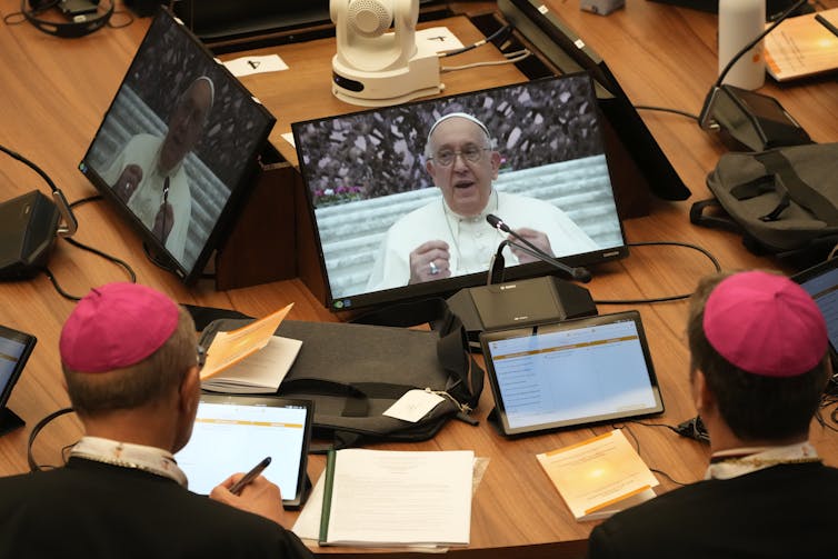 Two men with pink skullcaps sit at a table watching the pope speak on a large digital display propped on the table.