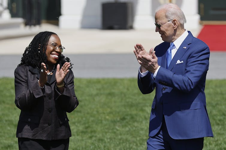 A woman in a black jacket and a man in a blue suit standing outside applaud.