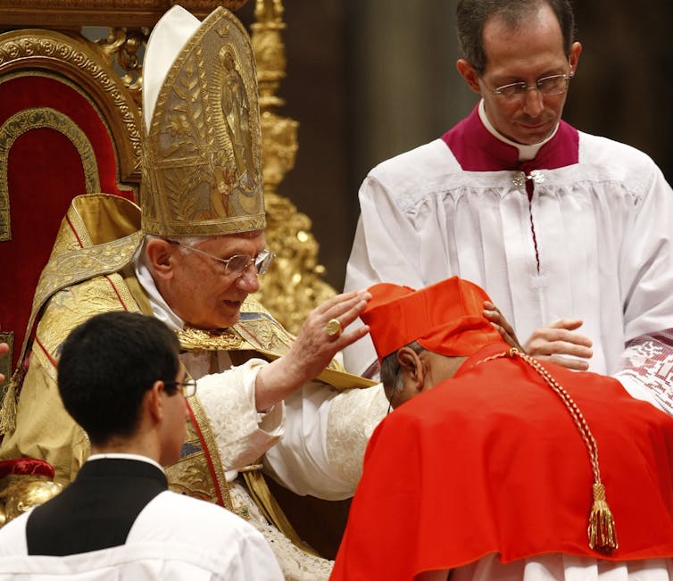 A man in a red head dress bows before the pope seated on a chair.