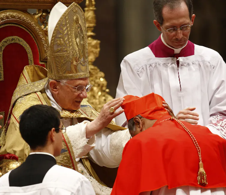 A man in a red head dress bows before the pope seated on a chair.