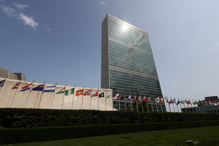 A tall glass building has the sun shine on it. A row of country flags surround the building at its base.