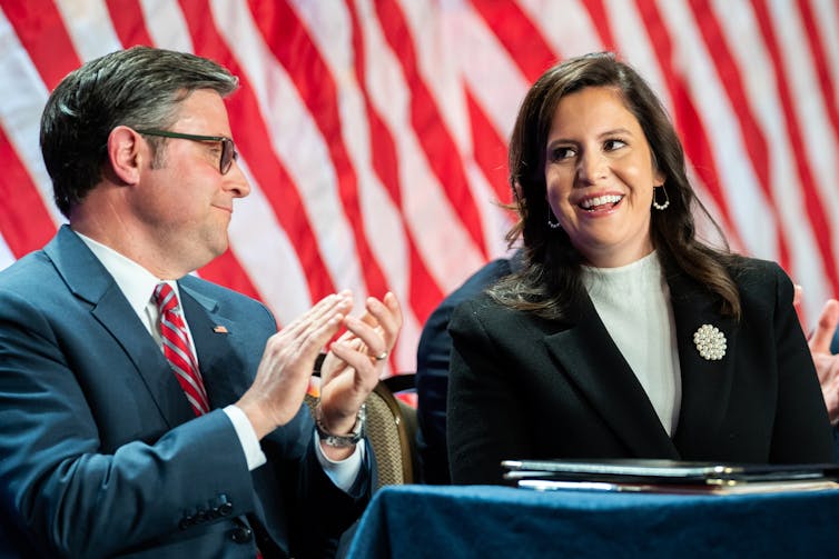 A woman with dark hair and a black blazer smiles in front of American flags. A man next to her claps and smiles.