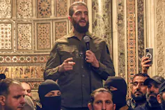 A thin bearded man in camouflage clothing speaks.