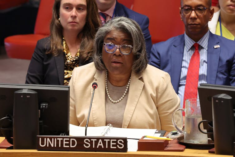 A brown skinned woman with a beige blazer and grey hair speaks into a microphone. She is seated at a desk with a placard that says 'United States'