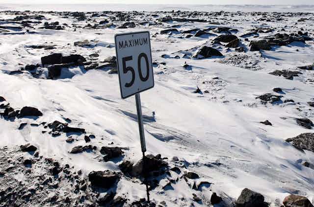 A speed limit sign is seen in a snowy, craggy expanse with no road visible.