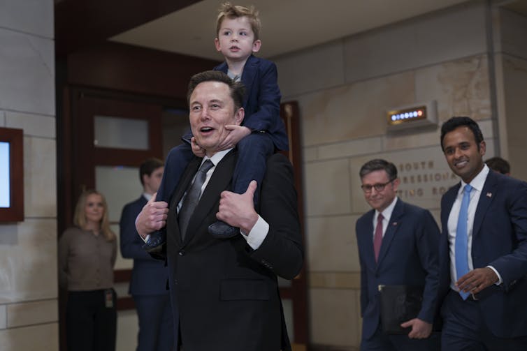 a young boy piggy backs on a man wearing a suit with two other men in suits smiling behind him