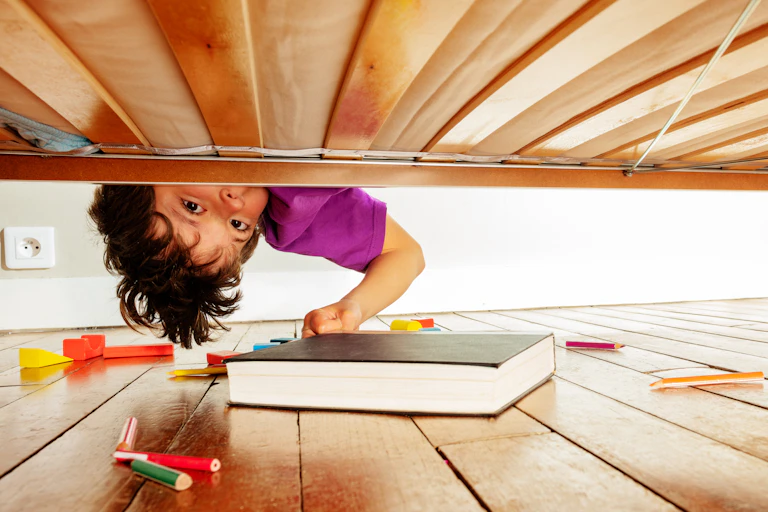 Child's face peering upside down under bed where books, pencils and blocks are lying.