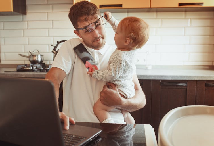 A toddler takes glasses and phone off a man working on a laptop.