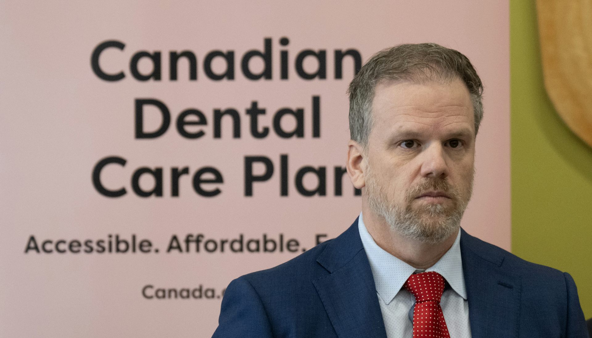 A man in a blue suit and red tie stands in front of a pink sign reading 'Canadian Dental Care Plan'