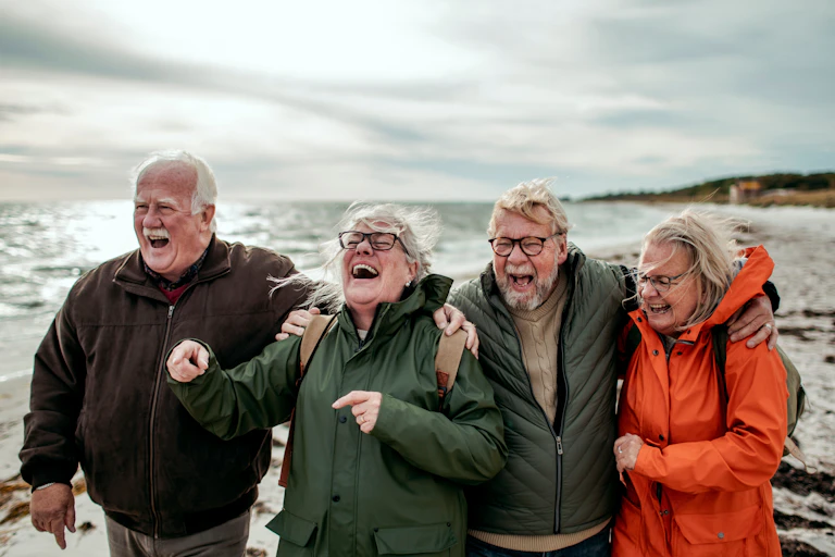 Un groupe de quatre retraités (deux hommes et deux femmes) qui marchent en riant au bord de la mer