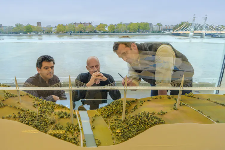 Three men look at model bridge. River in background