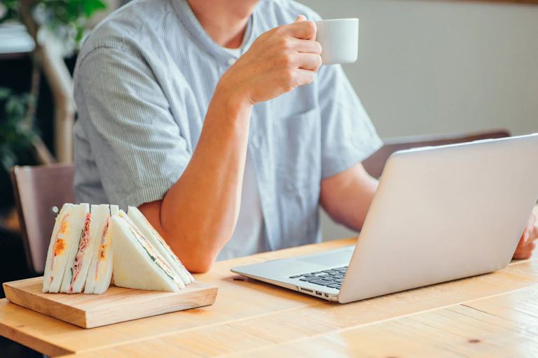 Un homme devant son ordinateur avec un café à la main et un sandwich prêt à etre mangé