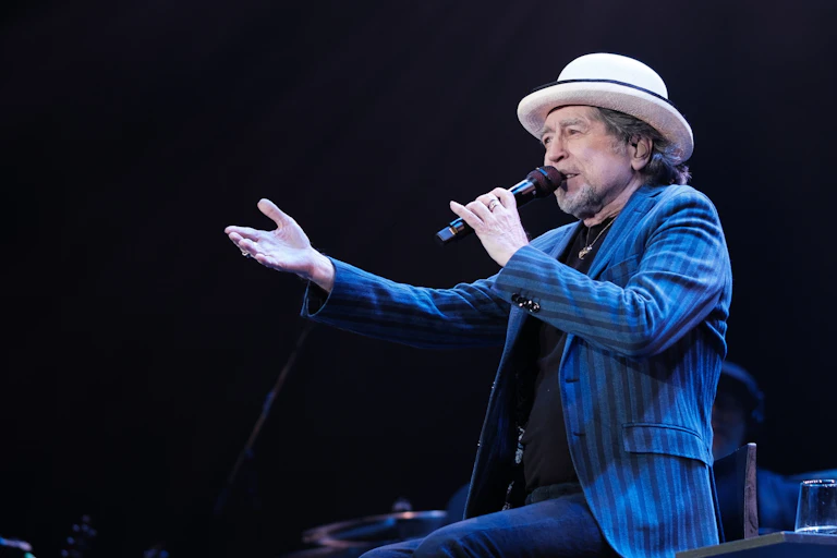 Un hombre con un sombrero y una americana azul canta sentado y señala al público.