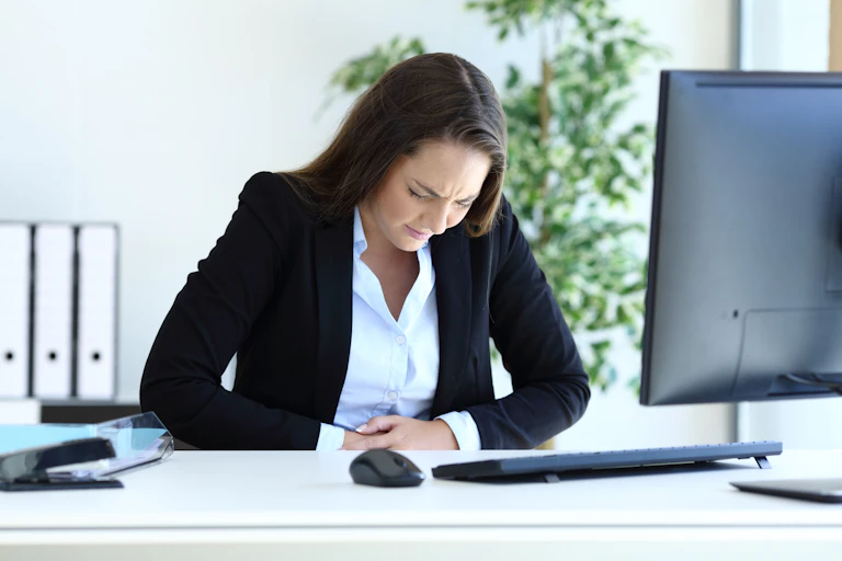 Une jeune femme se tient le ventre de douleur dans son bureau, face à son ordinateur professionnel.