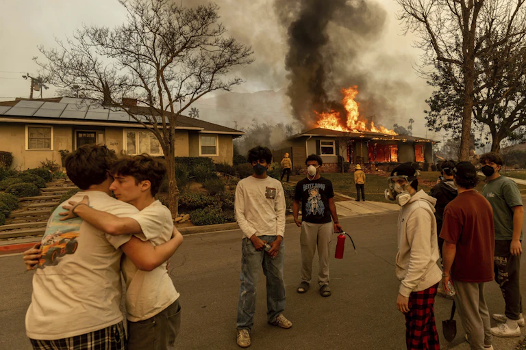 Two young people hug each other outside a home that is on fire. Dark smoke rises from the home and the sky is orange-gray.