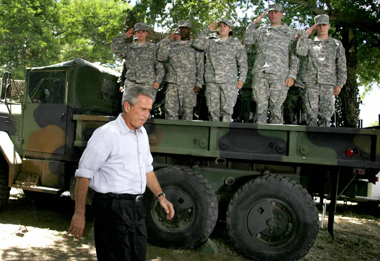 National Guard soldiers standing in the bed of a truck salute President George W. Bush as he walks past