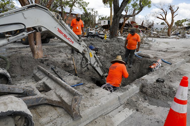 Three men in orange dig beneath a devastated street