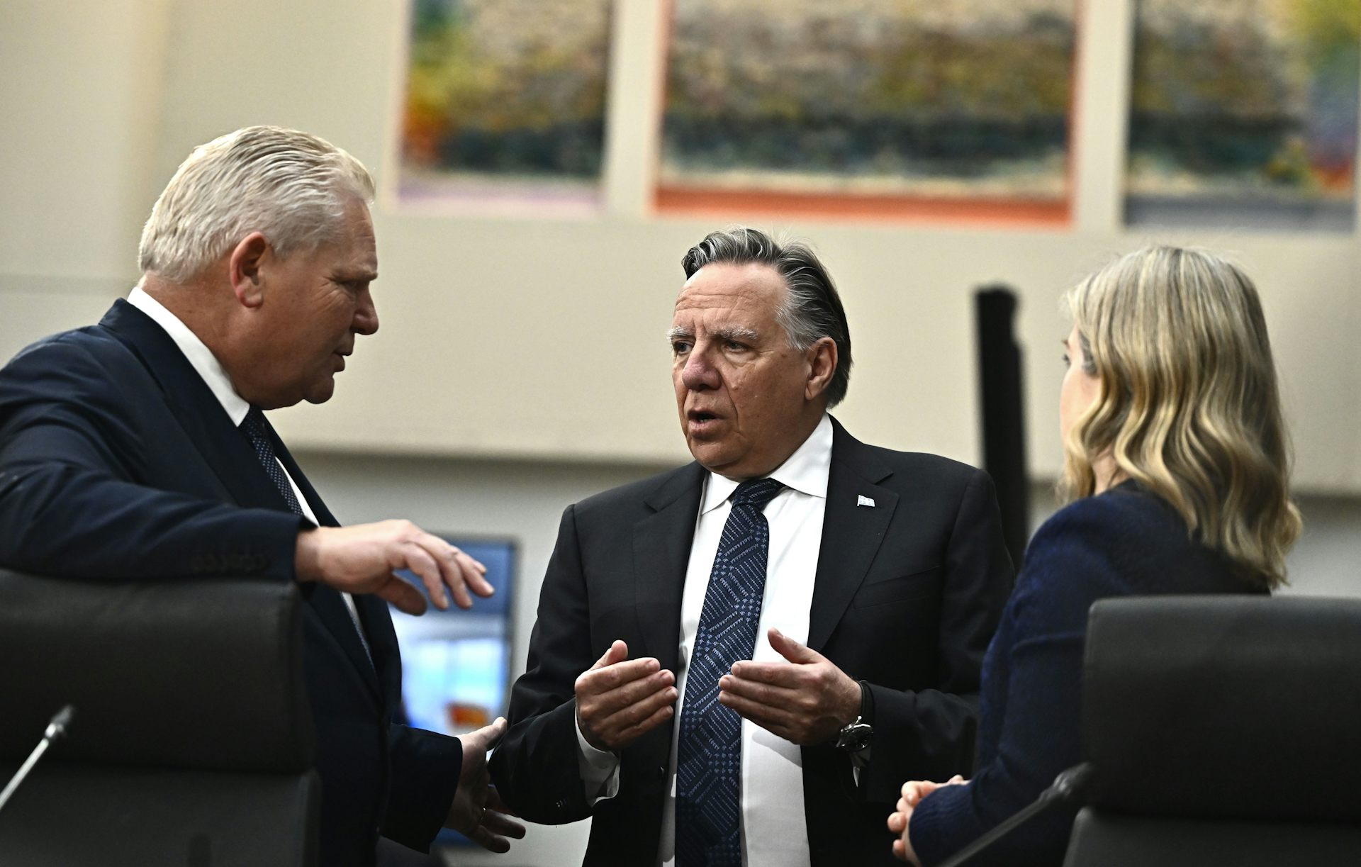 Two men in suits and a woman with blond hair stand talking.