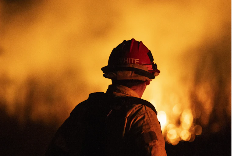 With his back to the camera, a firefighter looks at an enormous fire raging in the distance.