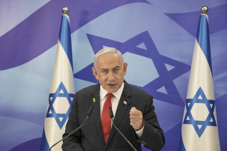 A man in a suit gives a speech at a lectern in front of two Israeli flags