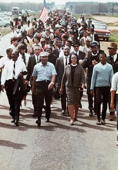 A crowd of people walking in lines march in suits and ties, with one participant holding an American flag.