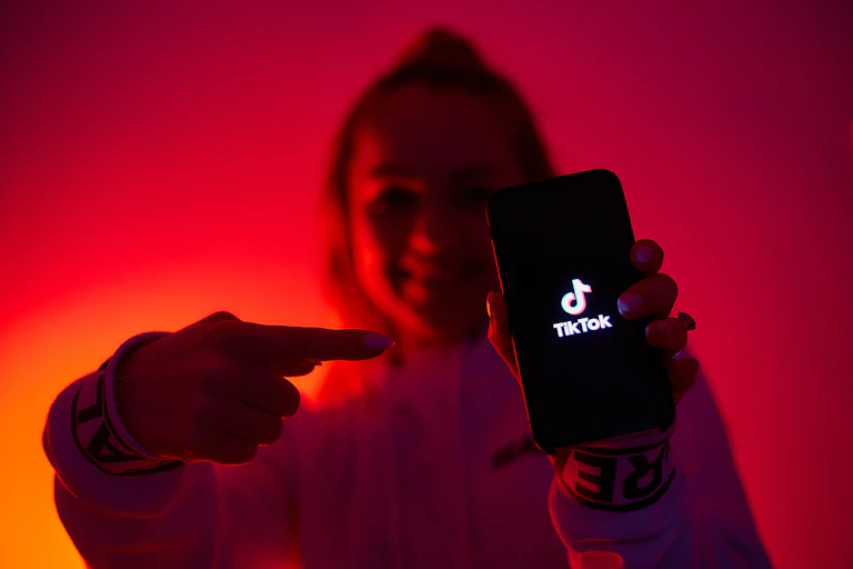 A girl standing against a glowing red and orange background, pointing to a phone displaying the TikTok logo.