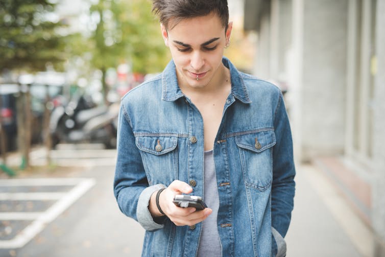 Young, pale man walking along street, looking down at phone in hand