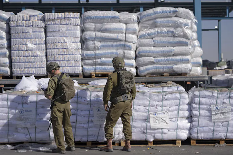 Dois soldados estão em frente a pilhas de sacos brancos cheios de materiais de ajuda